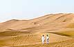 Two people walk across a sand dune.